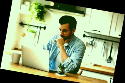 El hombre se sienta a la mesa en casa, cocina, trabajo en línea en el portátil, reflexionando sobre el pensamiento