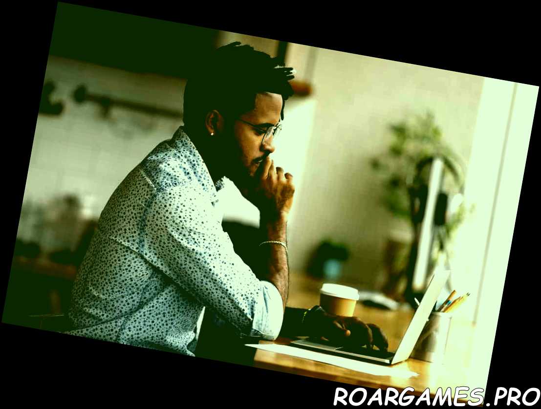 un joven birracial con anteojos se sienta en el escritorio y mira la pantalla de un portátil trabajando en línea en la oficina de casa